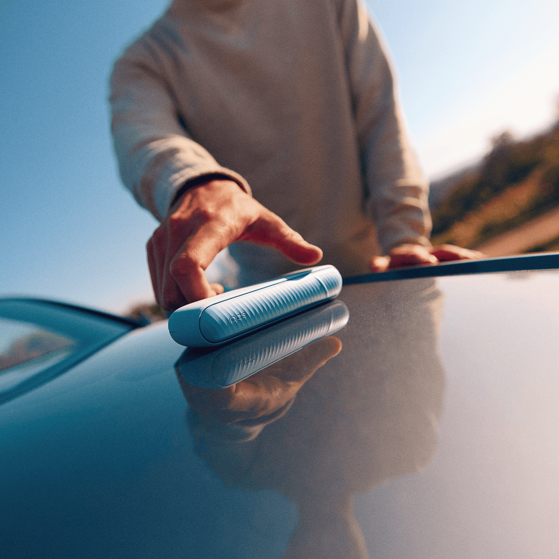 A man holding an IQOS ILUMA i color Breeze Blue on top of a car